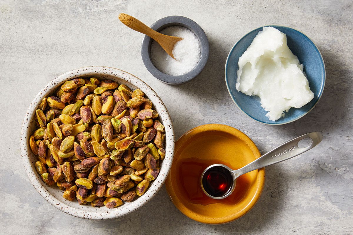 Overhead ingredient layout of toasted pistachios arranged beside melted coconut oil, vanilla extract and salt on a neutral surface. This image highlights the simple pantry ingredients used to create homemade Pistachio Butter.