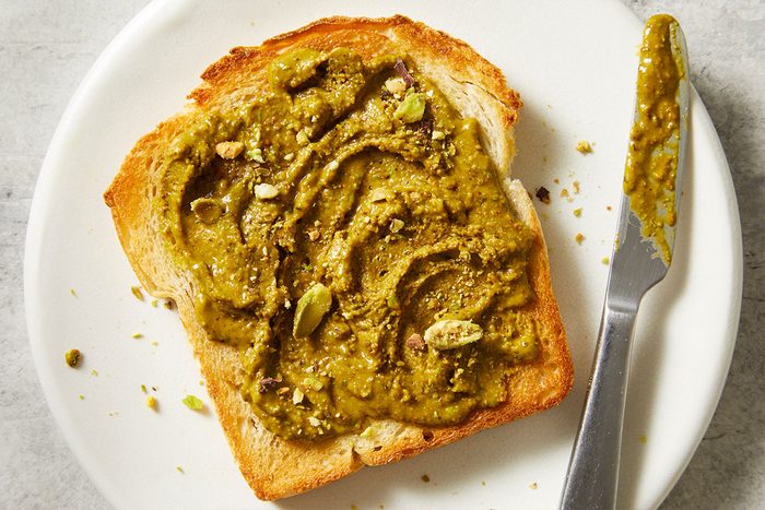 Horizontal overhead lifestyle image of Pistachio Butter generously spread on toasted bread. The textured swirl of green nut butter showcases its versatility as a breakfast or snack topping.