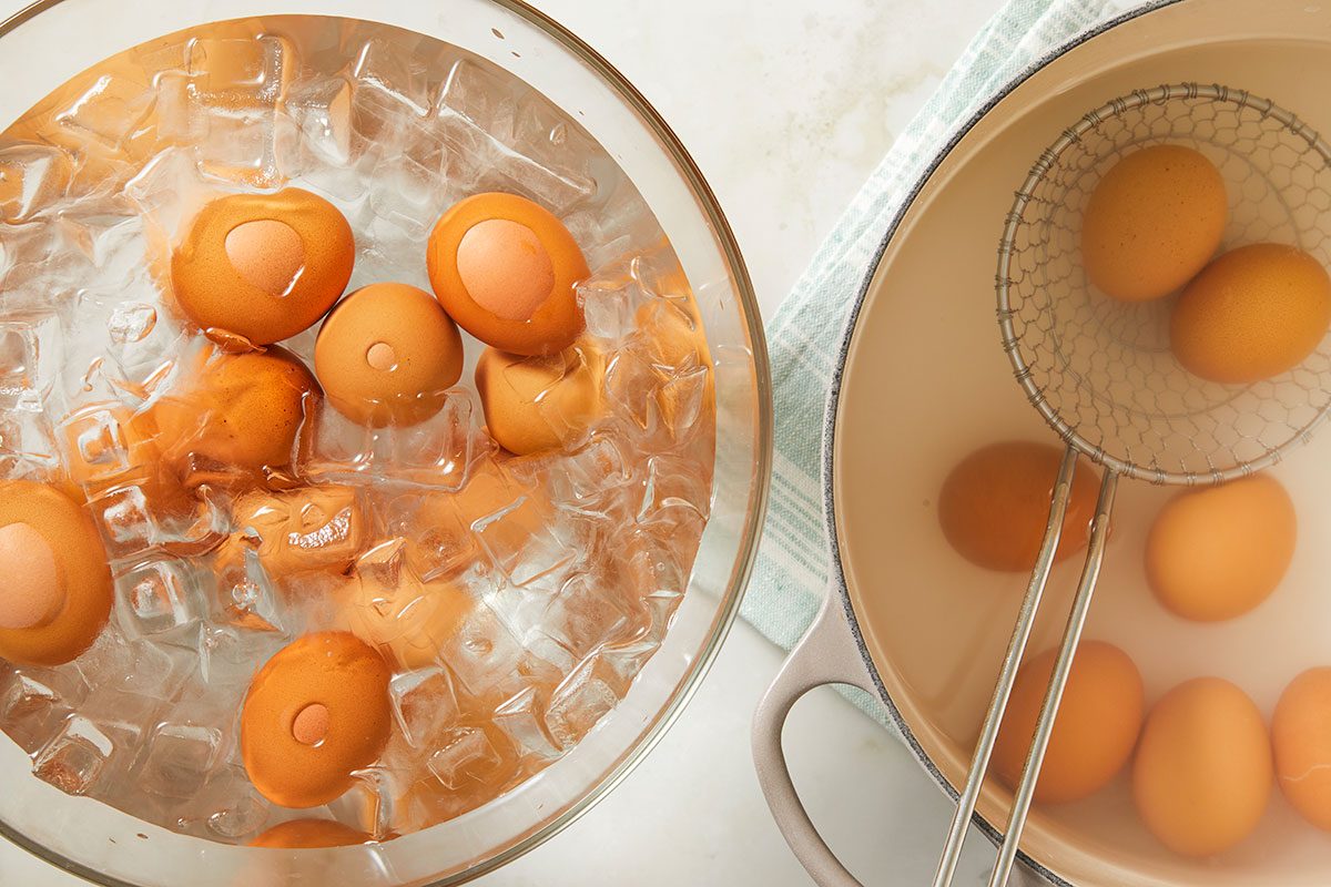 Overhead image of hard-boiled eggs resting in an ice bath alongside eggshells and utensils on a light countertop. This image illustrates the cooling step after boiling eggs for the deviled egg mixture.