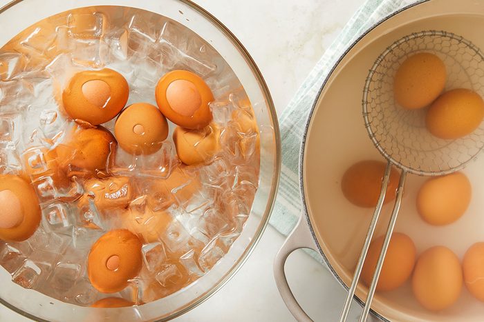Overhead image of hard-boiled eggs resting in an ice bath alongside eggshells and utensils on a light countertop. This image illustrates the cooling step after boiling eggs for the deviled egg mixture.