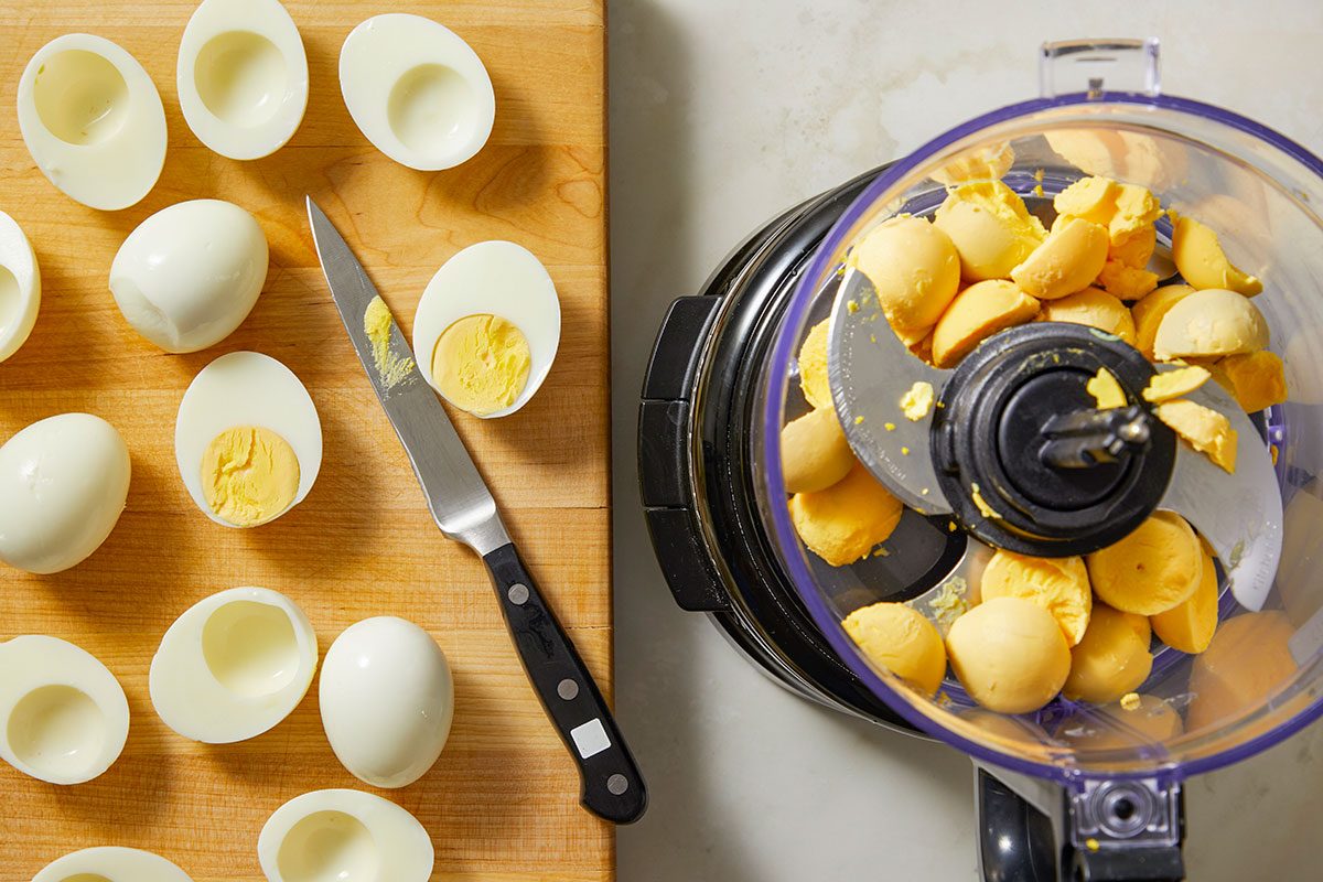 Top-down view of peeled hard-boiled eggs arranged on a cutting surface next to a knife, prepared for processing into deviled egg filling. The image highlights mise en place for a holiday appetizer.