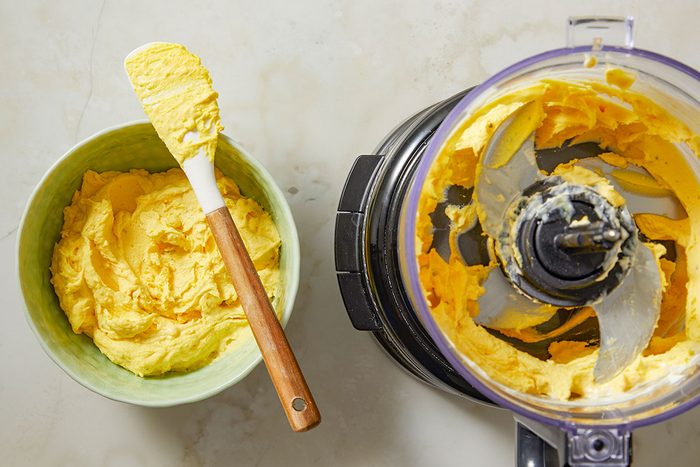 Overhead view of peeled eggs placed inside a food processor bowl, ready to be blended for deviled egg filling. A small bowl of mustard sits nearby on the work surface.