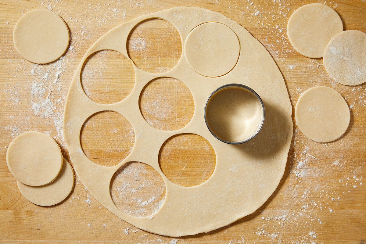Overhead step-by-step image showing refrigerated pie crust rolled out on a floured wooden surface with multiple circular cutouts removed using a biscuit cutter. Extra dough rounds are scattered around the board, demonstrating preparation for assembling a Pull-Apart Pie using refrigerated crust.