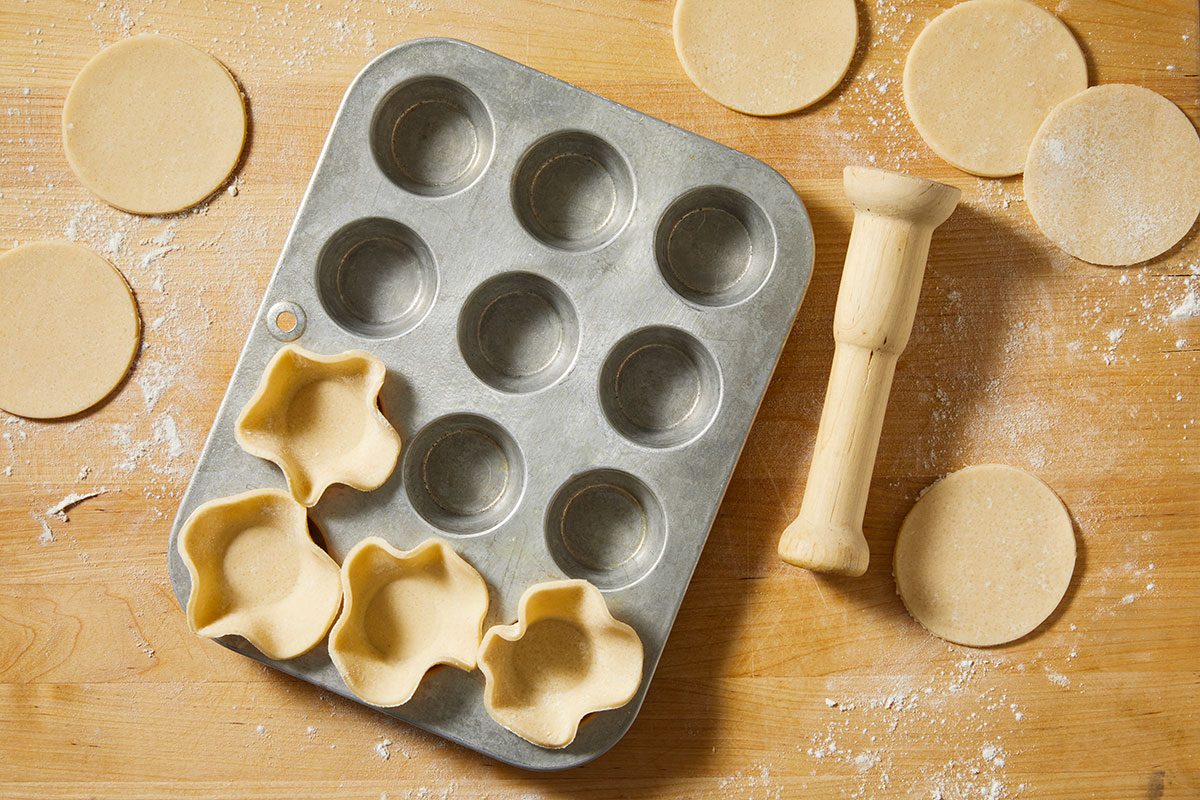 Overhead step-by-step image of pie crust circles pressed into a metal muffin tin to form individual pastry cups. A wooden tart tamper rests beside the pan on a lightly floured wooden surface, illustrating preparation for assembling Pull-Apart Pie portions.