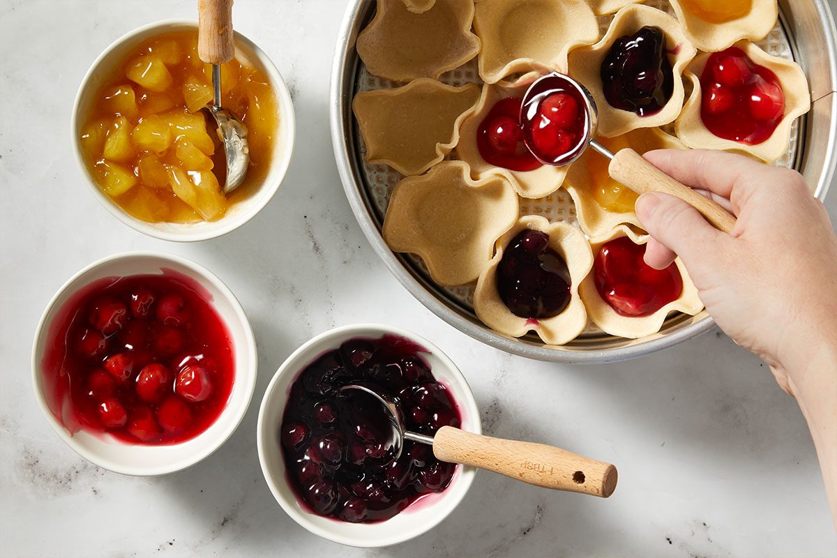 Overhead step-by-step image of mini pie crust cups arranged on a baking sheet while being filled with blueberry, cherry, and diced apple pie fillings. Bowls of fruit filling surround the pan as a hand spoons filling into the pastry shells, showing assembly of Pull-Apart Pie.