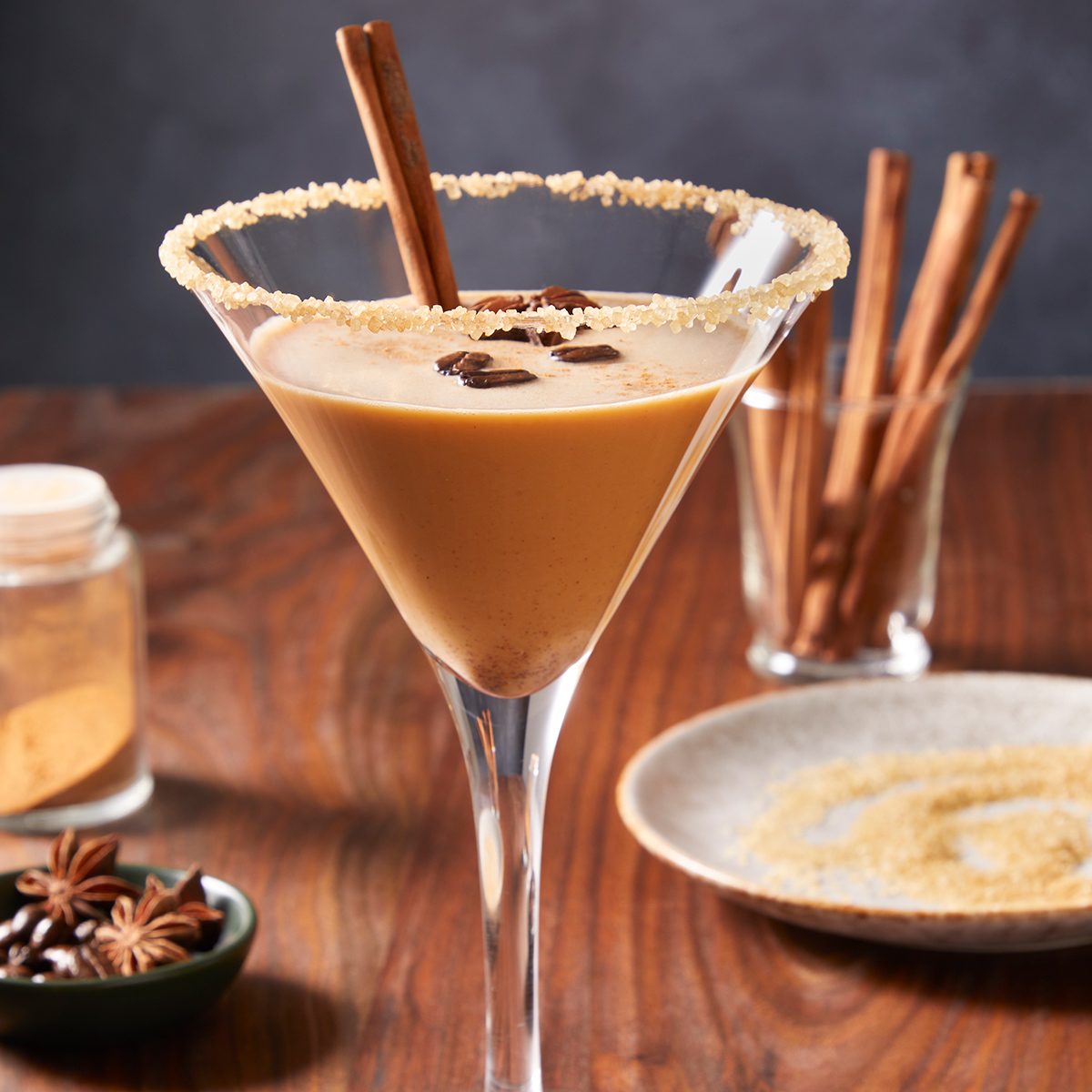 A creamy brown cocktail in a martini glass, rimmed with sugar, garnished with a cinnamon stick and coffee beans. There are spices and a jar in the background on a wooden table.