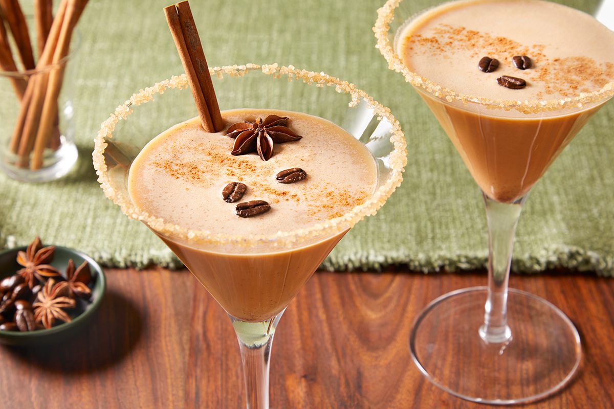 Two creamy coffee cocktails in martini glasses, garnished with coffee beans, star anise, and a cinnamon stick, with rimmed glasses and a green table mat underneath on a wooden surface.