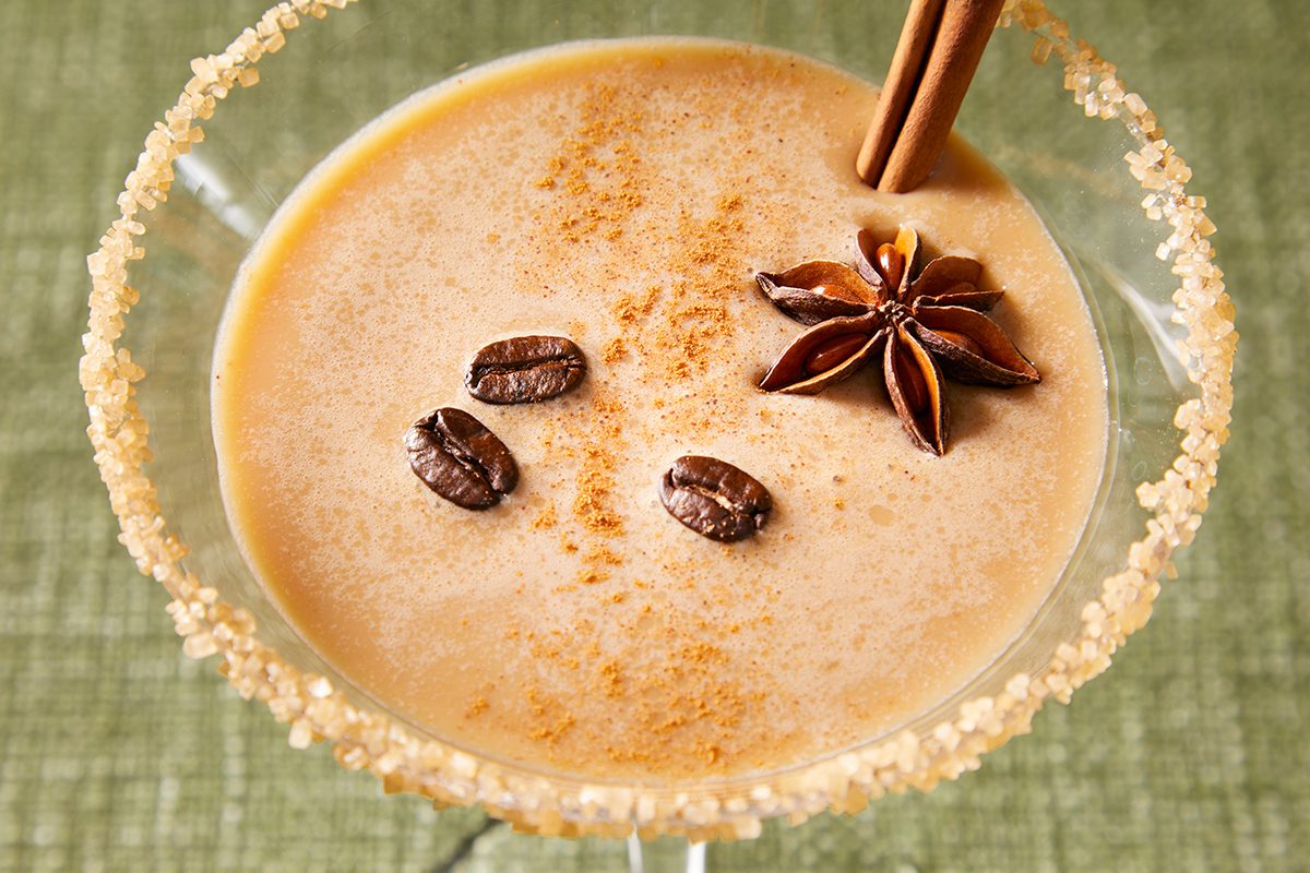 A creamy coffee cocktail in a rimmed glass, garnished with three coffee beans, a star anise, and two cinnamon sticks on top, set against a green textured background.