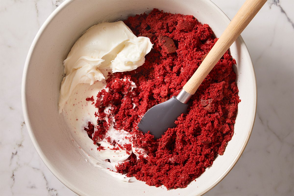 A overhead, horizontal shot of Red Velvet Cake bites being prepped from a bowl of cake batter, scooped with a round scoop—cake bite balls are then placed on the baking sheet