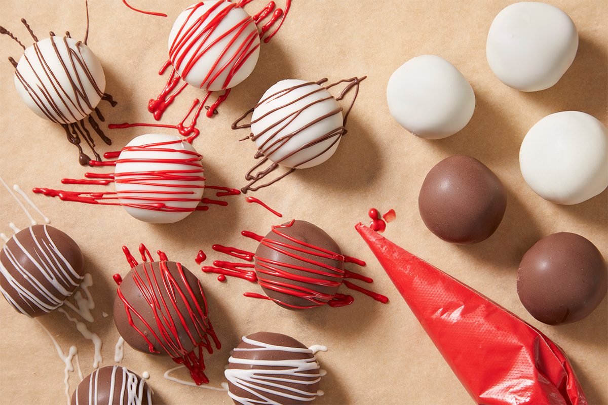 A overhead, horizontal shot of Red Velvet Cake bites on parchement paper being decorated with red frosting in a tube—some undecorated Cake Bites remain in the upper right of the image.