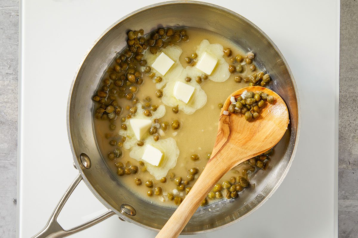 Overhead step-by-step image of butter melting in a skillet with minced garlic and capers beginning to sauté. A wooden spoon rests in the pan as the sauce base for Salmon Piccata is prepared.