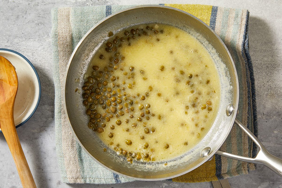 Overhead step-by-step image showing dry white wine and reduced-sodium chicken broth added to the skillet with capers and butter, forming the lemon piccata sauce.