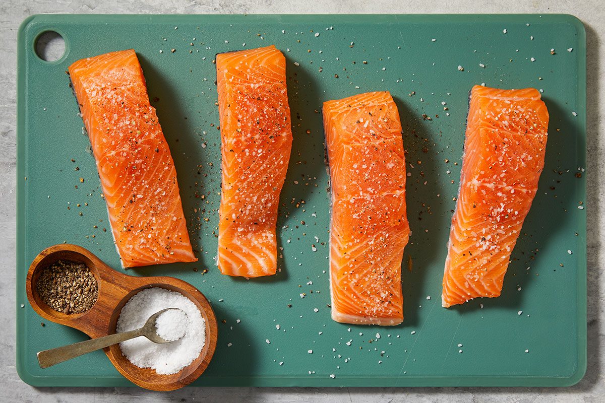 Overhead step-by-step image of raw salmon fillets arranged on a green cutting board, seasoned with salt and pepper, with small bowls of seasoning nearby before cooking.