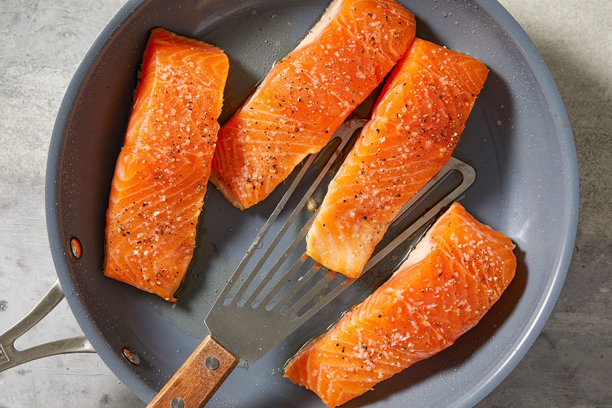 Overhead step-by-step image of salmon fillets searing in a skillet with olive oil. A spatula rests beside the fish as the fillets cook to develop a golden crust.
