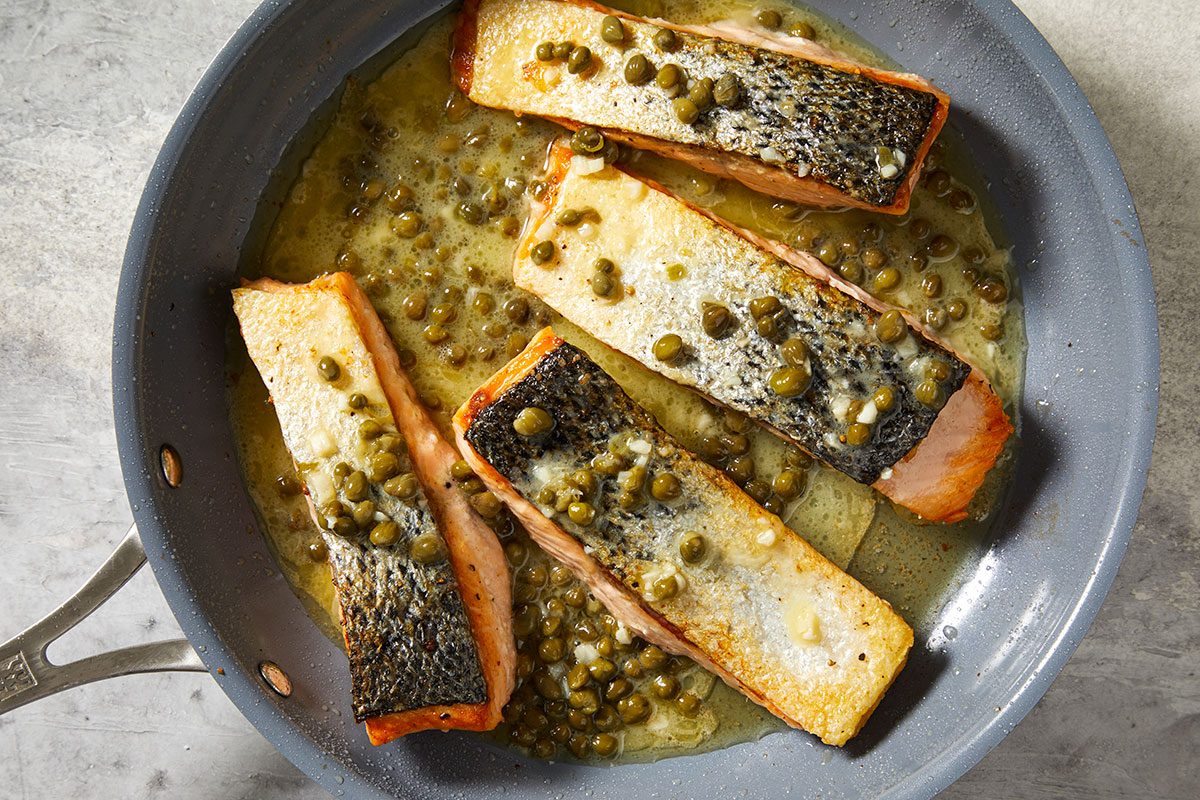 Overhead step-by-step image of seared salmon fillets nestled in lemon caper butter sauce inside the skillet, finishing cooking in the flavorful piccata sauce.