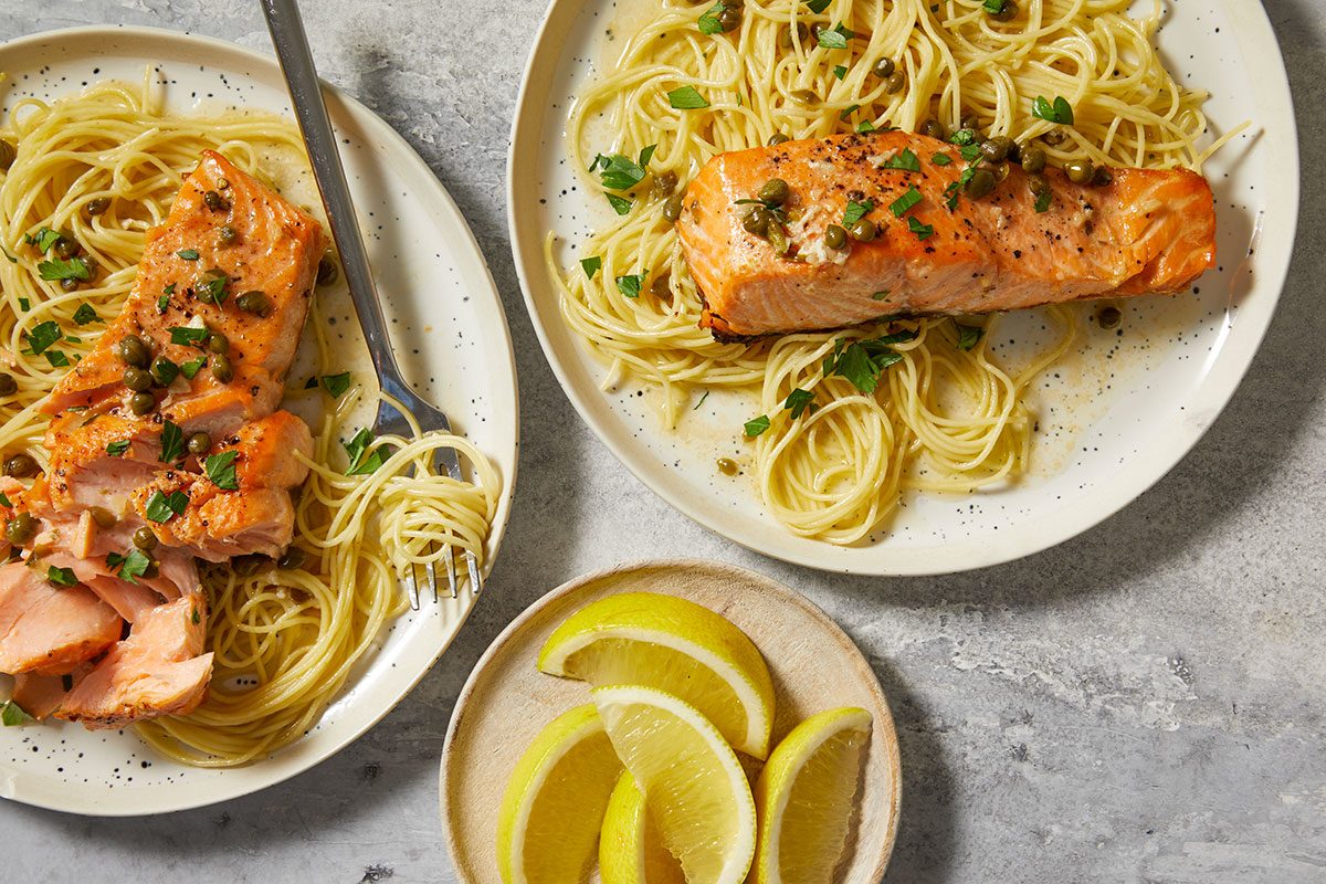 Overhead Push and Pulls image of Salmon Piccata served over a bed of pasta, topped with lemon caper sauce and garnished with parsley, with lemon wedges on the side.