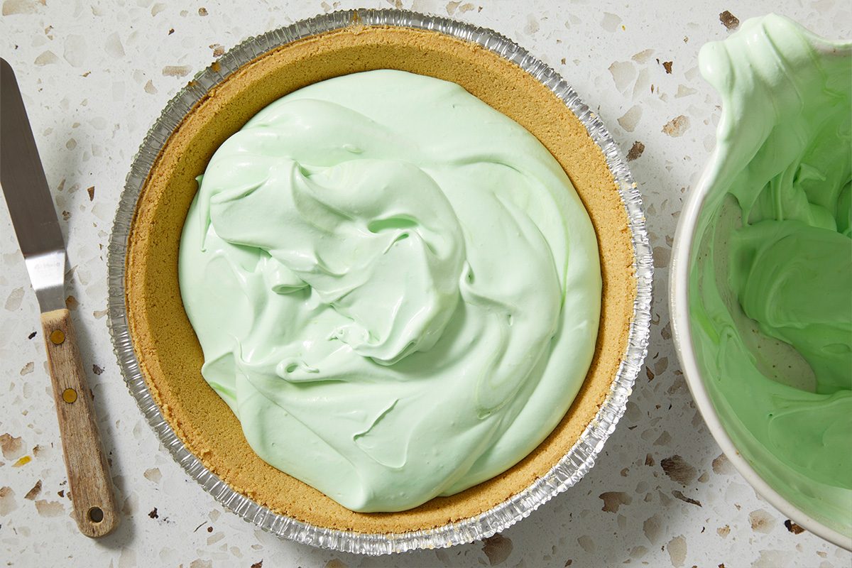 Overhead, horizontal image of the green Shamrock Shake Pie filling spread evenly inside a graham-style pie crust. A spatula rests nearby, highlighting the assembly stage before chilling or setting.
