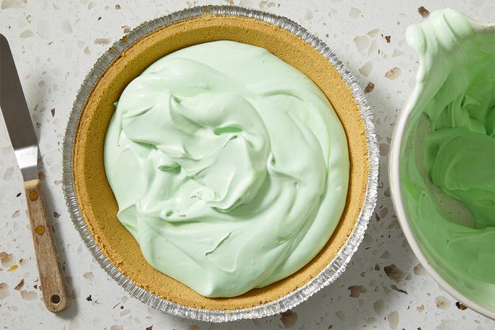 Overhead, horizontal image of the green Shamrock Shake Pie filling spread evenly inside a graham-style pie crust. A spatula rests nearby, highlighting the assembly stage before chilling or setting.