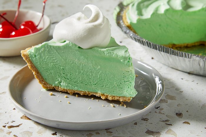 Overhead, horizontal close-up of a single slice of Shamrock Shake Pie served on a plate. The green filling is topped with whipped cream and a red cherry, emphasizing contrast and texture.