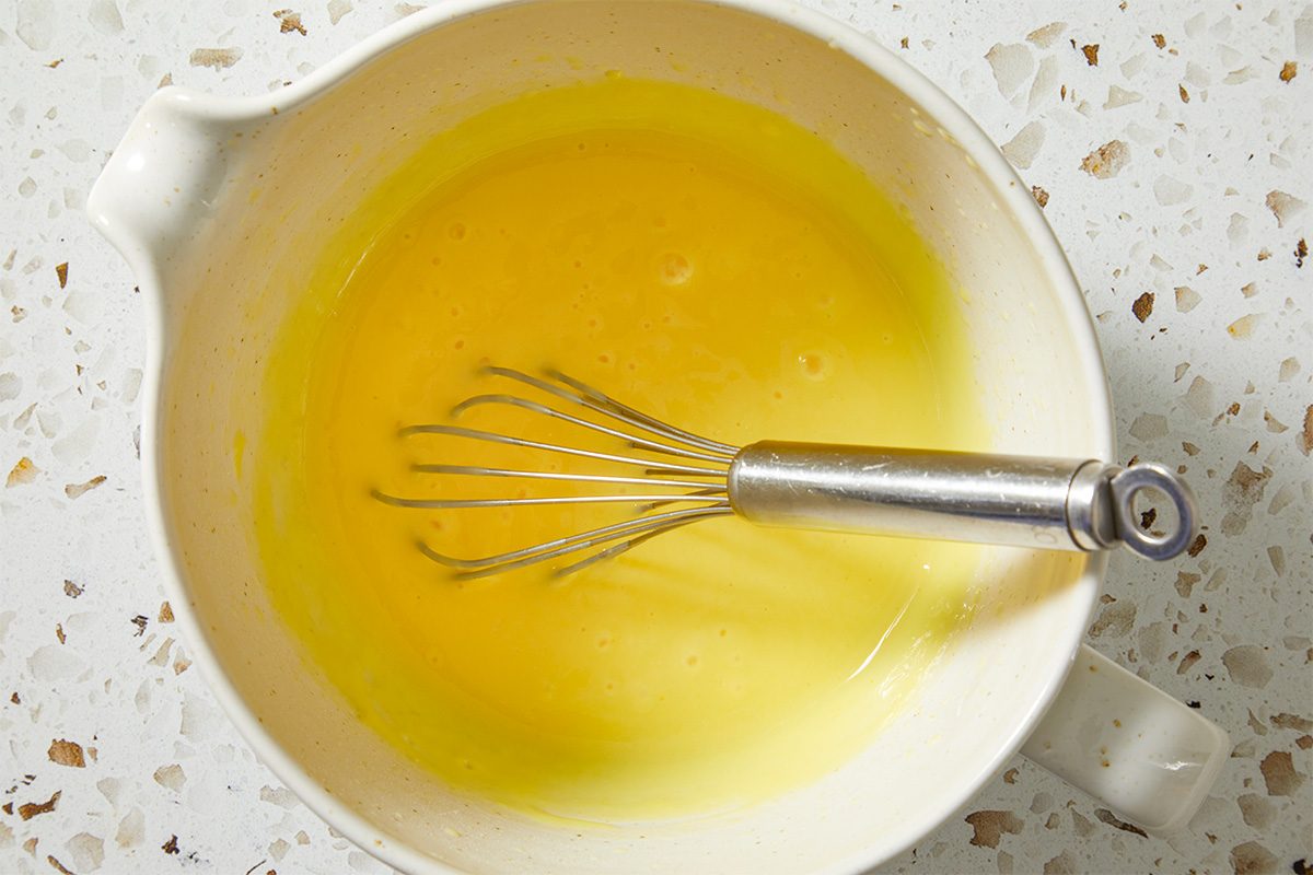 Overhead, horizontal step-by-step image showing a yellow liquid mixture being whisked in a bowl. A metal whisk rests in the center as the smooth base is combined, representing the early preparation stage of Shamrock Shake Pie.