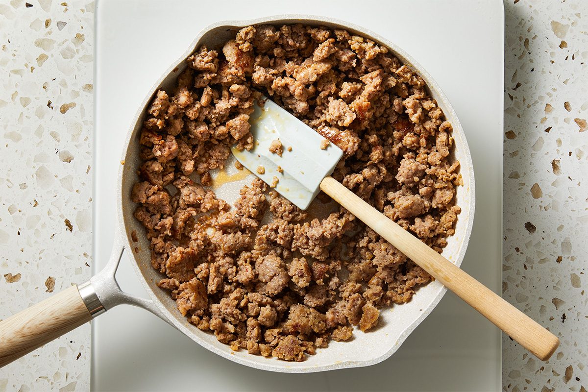 Overhead shot of browned and crumbled sausage cooking in a white skillet, with a spatula resting inside as the meat finishes sautéing on a speckled countertop.