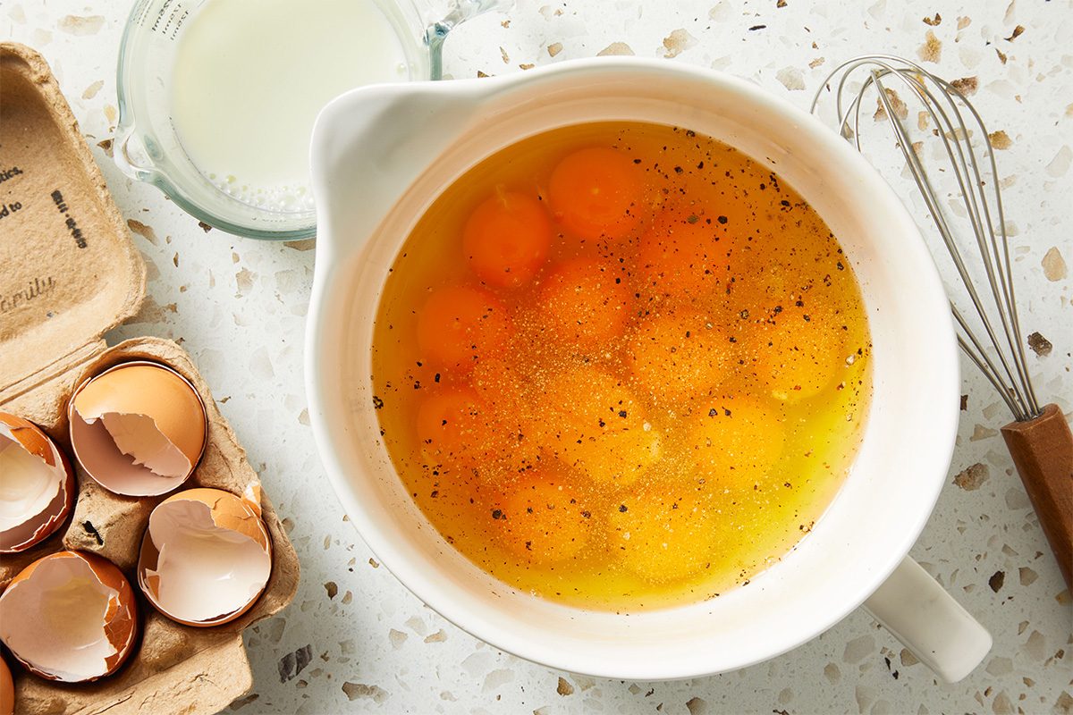 Overhead shot of a mixing bowl filled with cracked eggs, surrounded by broken eggshells and a whisk, ready to be poured into the slow cooker mixture. {