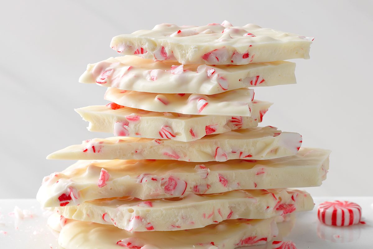 A stack of white chocolate bark pieces topped with crushed peppermint candy, with a whole peppermint candy visible next to the stack. The background is plain and light-colored.