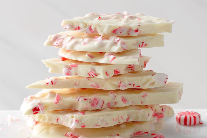 A stack of white chocolate bark pieces topped with crushed peppermint candy, with a whole peppermint candy visible next to the stack. The background is plain and light-colored.