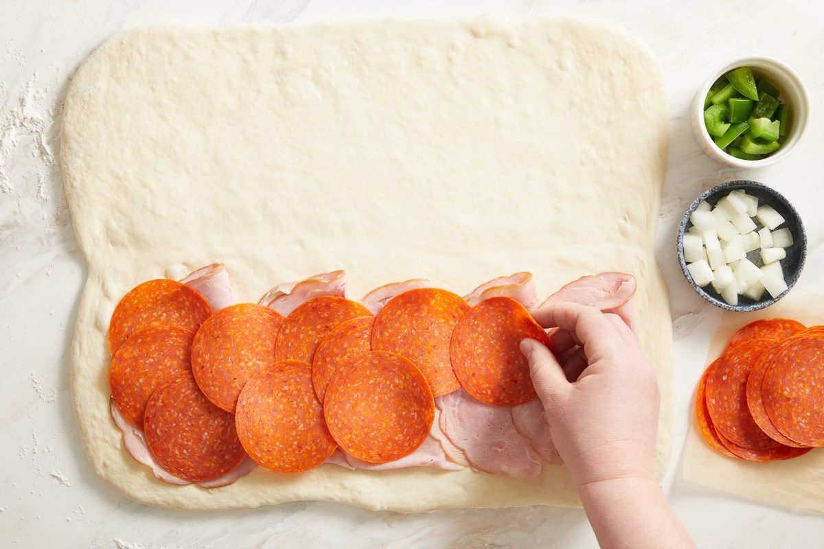 ham and pepperoni being layered on half of the dough lengthwise