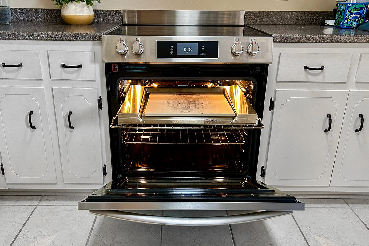 A stainless steel oven with its door open, showing a baking tray inside. The oven is built into white kitchen cabinets and sits below a gray countertop with various kitchen items on it.