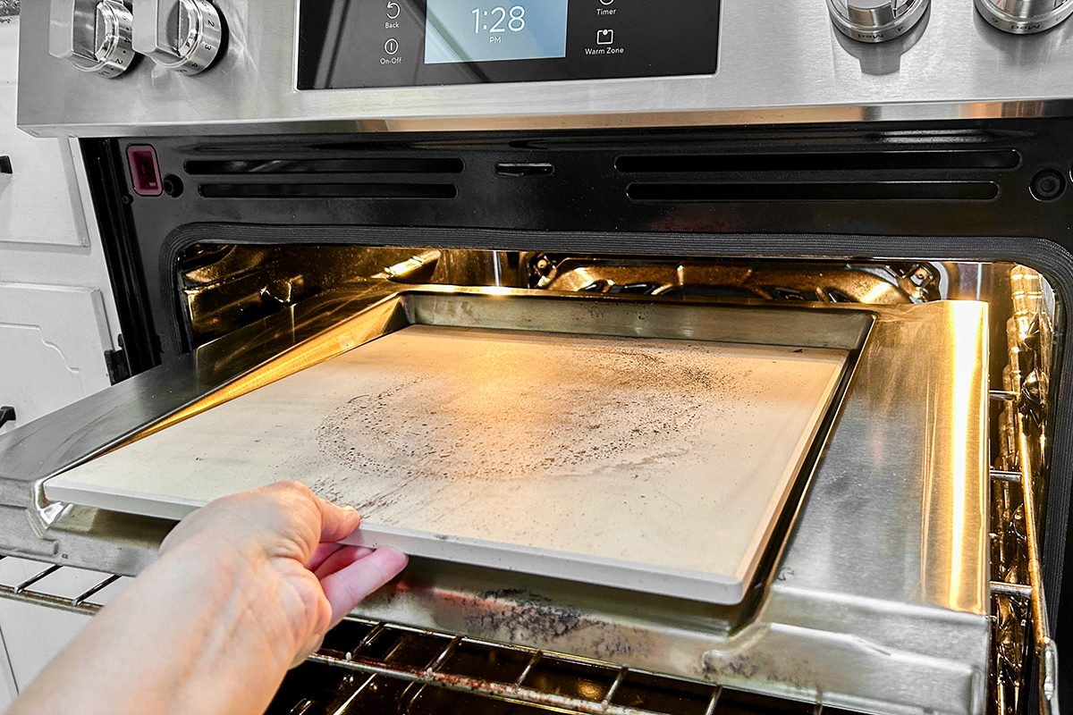 A hand is placing or removing a rectangular pizza stone from an open oven, with the stone resting on a metal baking tray inside the oven. The oven display shows the time "1:28.