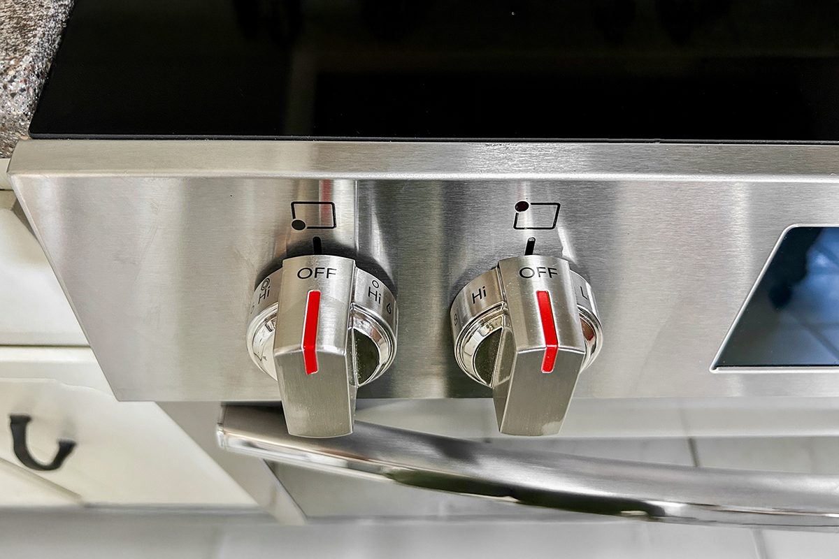 Close-up of two stainless steel stove knobs with red indicators, both pointed to the "off" position, on a modern oven with a sleek handle visible below.