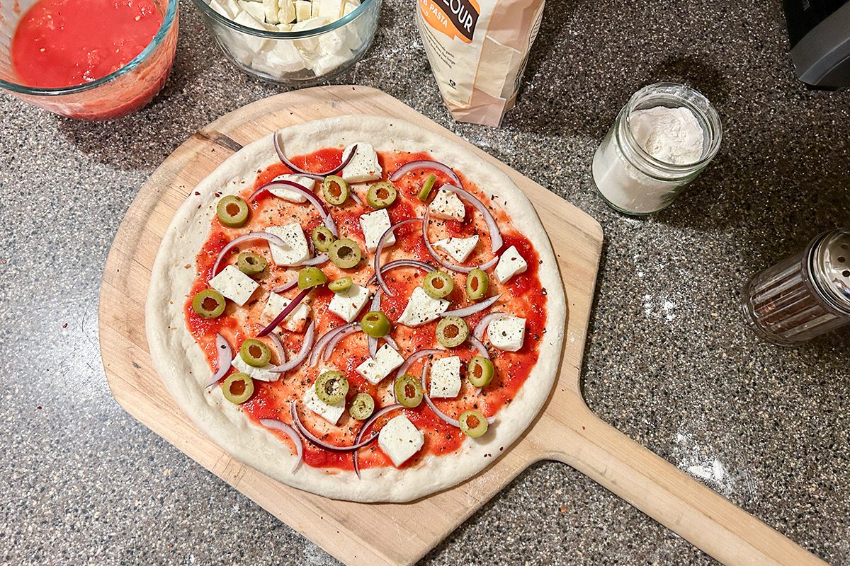 A homemade pizza on a wooden paddle, topped with tomato sauce, feta cheese cubes, green olives, and red onion slices. Ingredients like flour, cheese, and tomato sauce are on a granite countertop nearby.