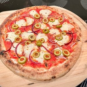 A freshly baked pizza on a wooden paddle, topped with mozzarella cheese, tomato sauce, green olives, and red onion slices, placed on a black surface with white circular patterns.