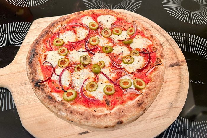 A freshly baked pizza on a wooden paddle, topped with mozzarella cheese, tomato sauce, green olives, and red onion slices, placed on a black surface with white circular patterns.