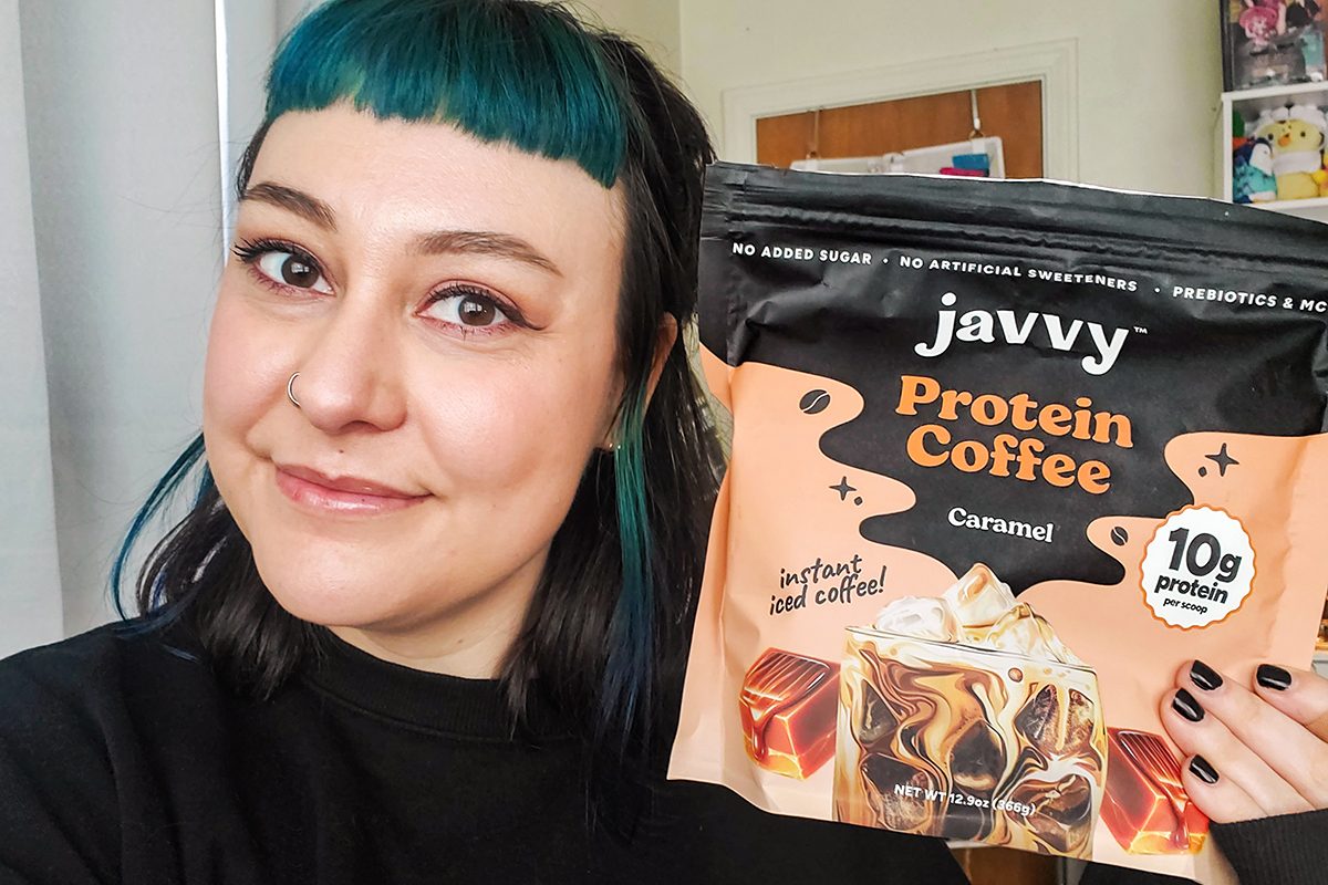 A person with teal and black hair smiles while holding a bag of Javvy Protein Coffee Caramel flavor, which features an image of iced coffee and caramel pieces on the packaging.