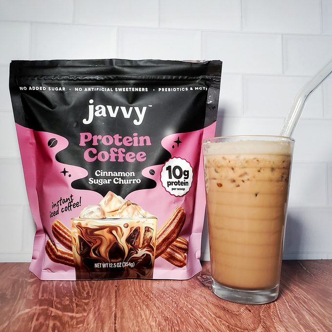 A bag of Javvy Protein Coffee in Cinnamon Sugar Churro flavor stands next to a glass of iced coffee with a clear straw, set on a wooden surface against a white tiled background.
