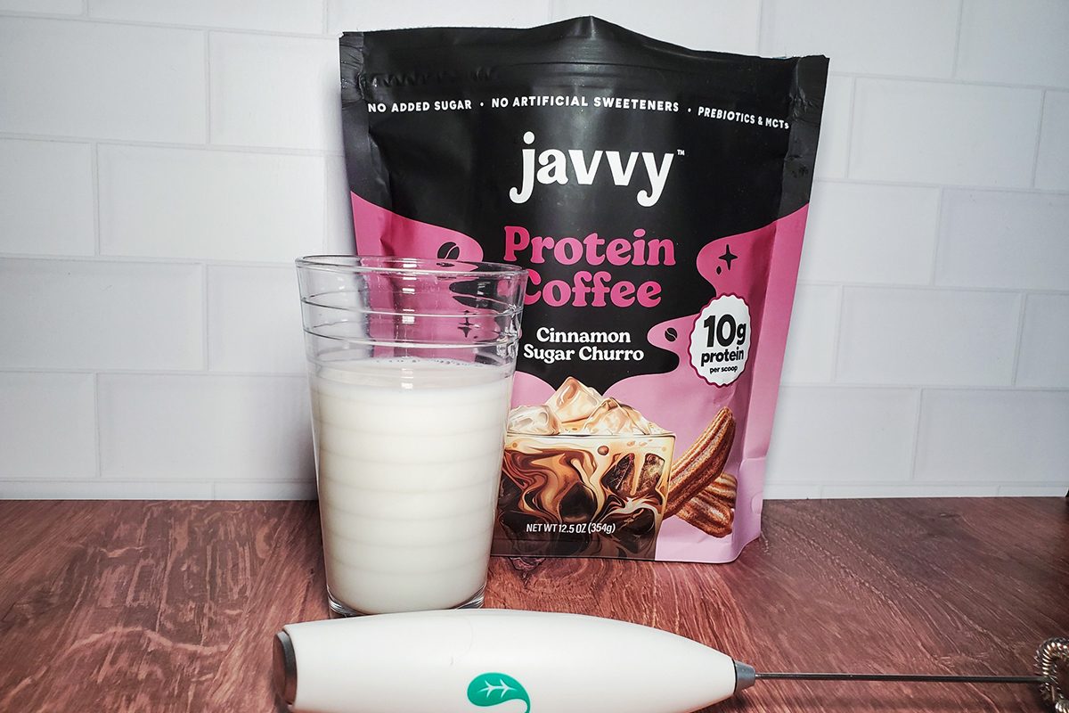 A glass of milk sits in front of a bag of Javvy Protein Coffee Cinnamon Sugar Churro flavor, with a handheld milk frother lying on a wooden surface nearby. The background shows white subway tile.