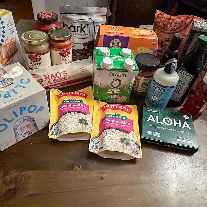 A variety of groceries on a table, including Rao’s pasta sauce, barkTHINS chocolate, Olipop soda, Tasty Bite rice packs, Orgain protein shakes, Aloha protein bars, and several canned and bottled items.