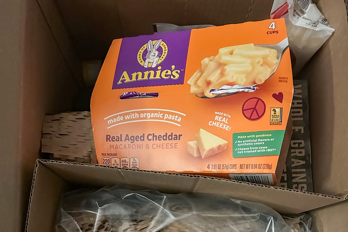 A box of Annie's Real Aged Cheddar Macaroni & Cheese sits inside a cardboard shipping box with other packaged items partially visible around it.