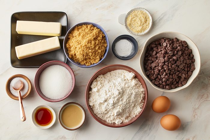 Assorted baking ingredients on a countertop, including sticks of butter, brown sugar, flour, chocolate chips, eggs, granulated sugar, vanilla extract, salt, baking powder, and a small bowl of milk.