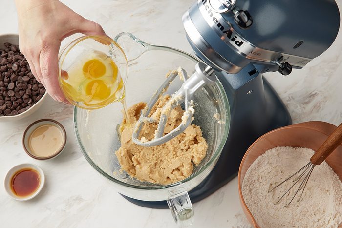 A person pours eggs into a stand mixer bowl with cookie dough. Nearby are bowls of flour with a whisk, chocolate chips, vanilla extract, and another ingredient on a white countertop.