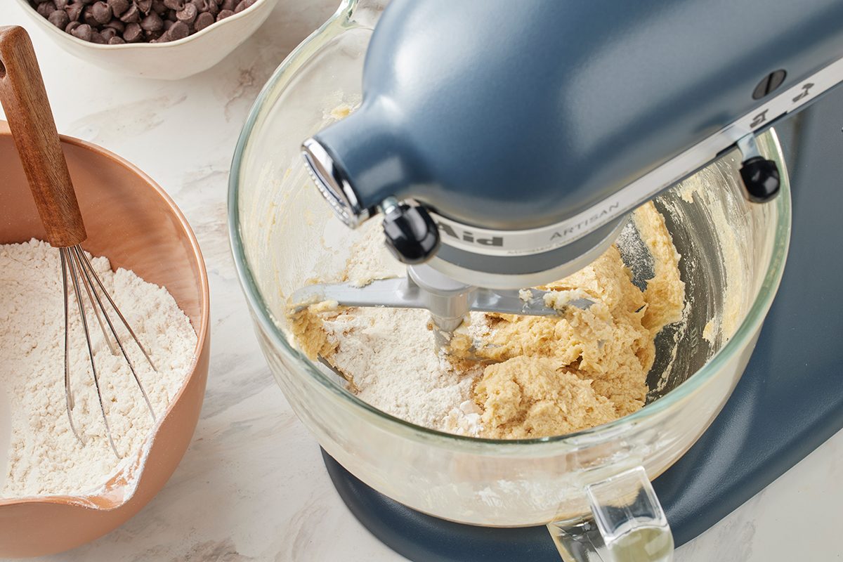 A stand mixer with a glass bowl is mixing cookie dough. A bowl of flour with a whisk and a bowl of chocolate chips are nearby on a light countertop.
