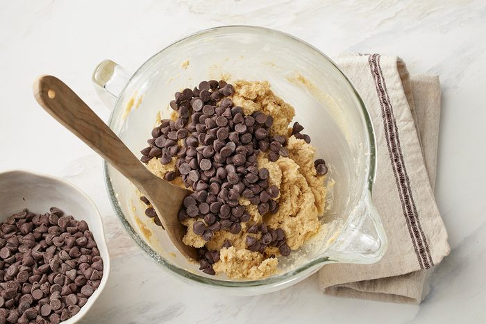 A glass mixing bowl filled with cookie dough and chocolate chips, being stirred with a wooden spoon, sits on a folded kitchen towel next to a bowl of more chocolate chips.