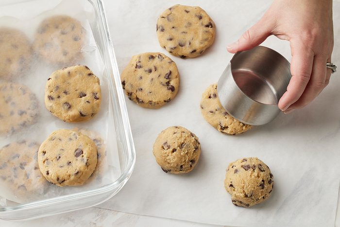 A hand uses a metal ring to shape chocolate chip cookie dough into round discs on parchment paper, with more dough shapes and a glass baking dish nearby.