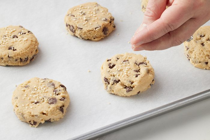 A hand sprinkles seeds on top of unbaked chocolate chip cookies arranged on a parchment-lined baking sheet.