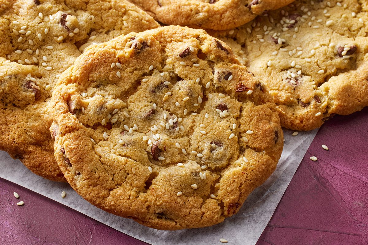 Close-up of several chocolate chip cookies with a golden-brown color and sprinkled with sesame seeds, arranged on a piece of parchment paper on a mauve surface.