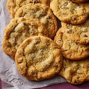A close-up of several chocolate chip cookies sprinkled with sesame seeds, arranged on a piece of parchment paper. The cookies look golden brown and freshly baked.