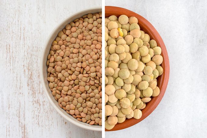 A side-by-side comparison of two bowls: the left bowl contains brown lentils, and the right bowl contains green lentils, both viewed from above on a light background.